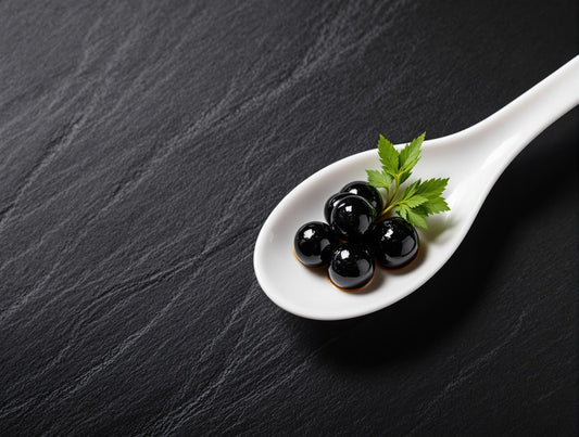 Dark, glossy Sticky Balsamic pearls the caviar alternative elegantly presented on a white ceramic spoon against a slate background, showcasing the rich black color and perfect 5mm spherical shape of premium balsamic vinegar pearls