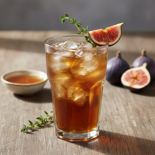 The image shows a tall glass of iced tea in warm amber tones, filled with ice cubes and garnished with a fresh green thyme sprig. A slice of fresh fig sits on the rim of the glass.