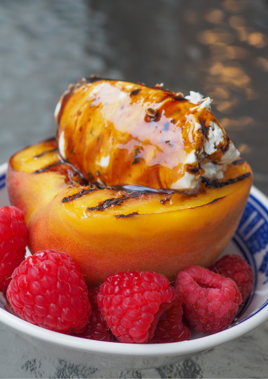 Grilled peach dessert drizzled with Sticky Balsamic, topped with creamy mascarpone ice cream and fresh red raspberries on a blue and white plate, close-up photo showing caramelized grill marks