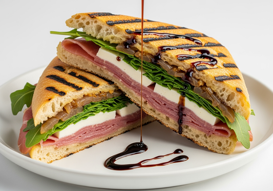 Golden-brown grilled panini cut in half showing layers of prosciutto, mozzarella, arugula, and caramelized onions with Sticky Balsamic Premium Fig drizzle