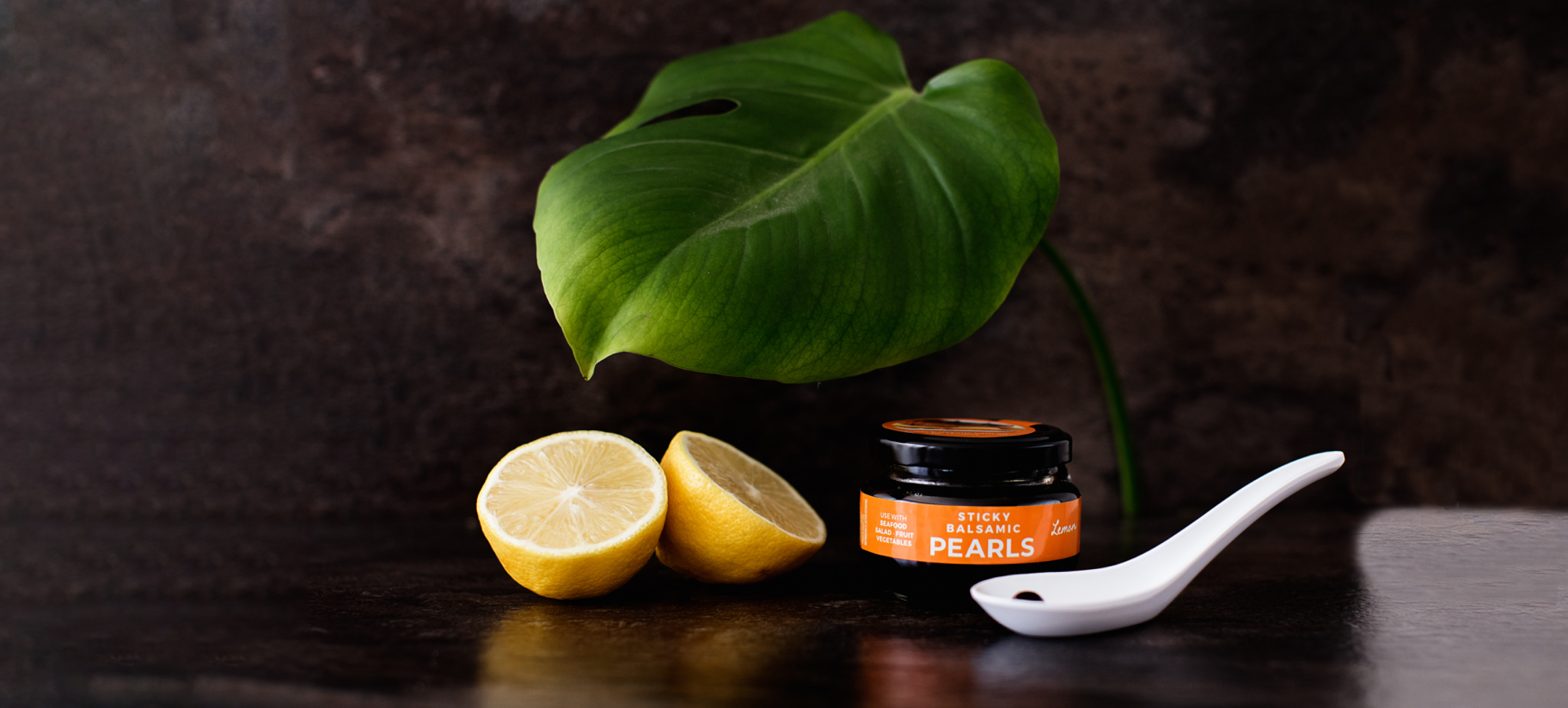 A jar of Sticky Balsamic Lemon Pearls with two half lemons, and a white spoon with a single Sticky Balsamic Pearl, with a leaf and dark background.