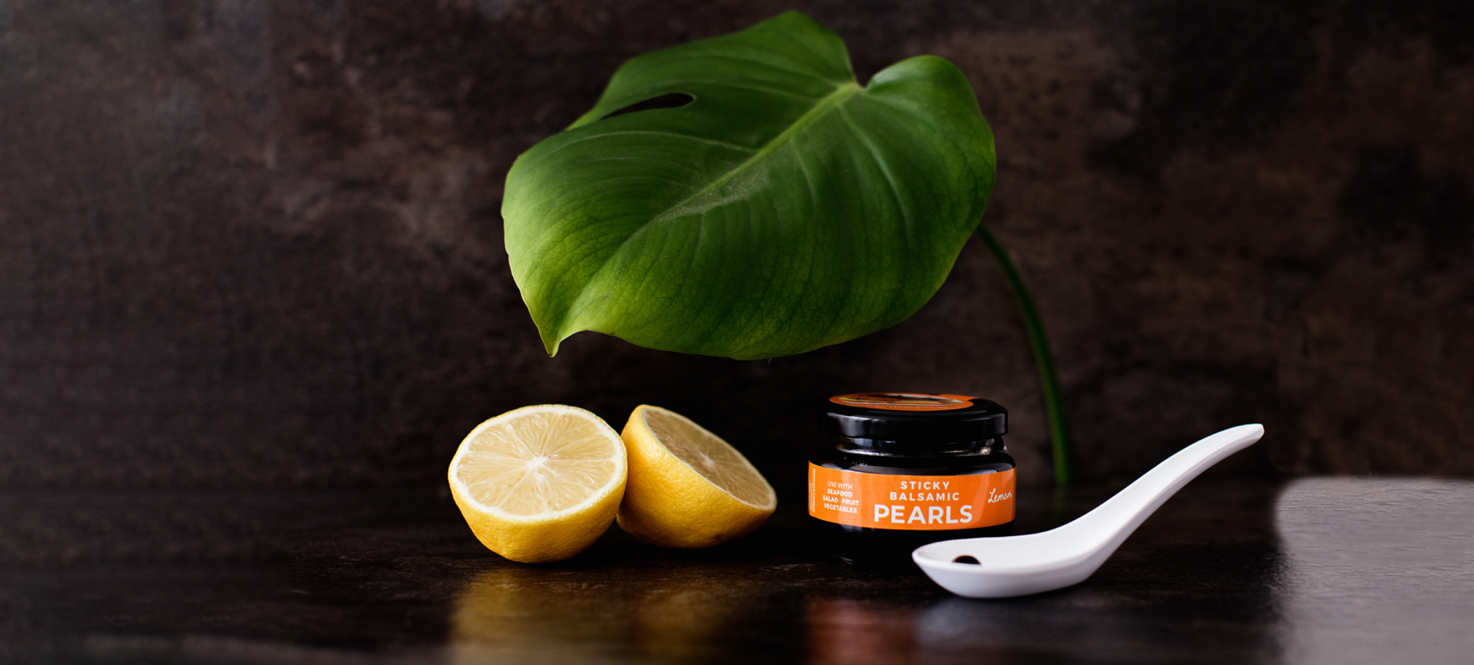 A jar of Sticky Balsamic Lemon Pearls with two half lemons, and a white spoon with a single Sticky Balsamic Pearl, with a leaf and dark background. Balsamic Vinegar