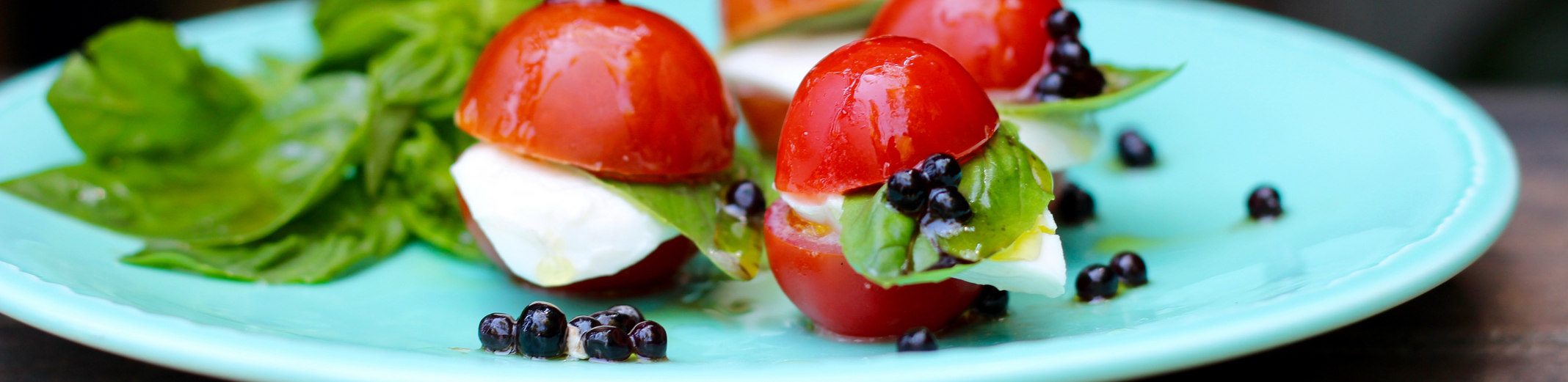 Caprese salad using mini tomatoes, a slice of buffalo mozzarella, basil and Sticky Balsamic pearls on a blue plate and wooden table.
