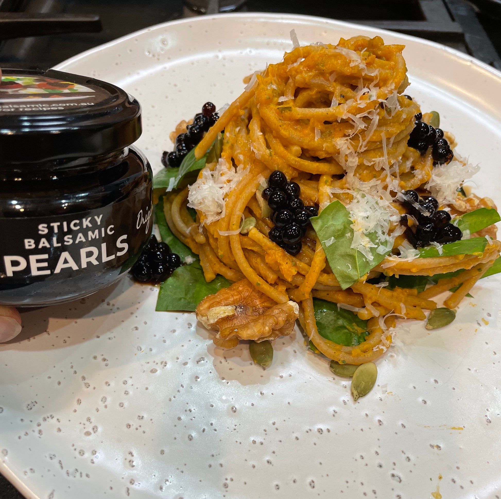 Easy Pumpkin Pasta with Sticky Balsamic Pearls  with spinach, walnuts and pepitas and grated parmesan on a white plate, with a jar of Sticky Balsamic Original Pearls on the side.