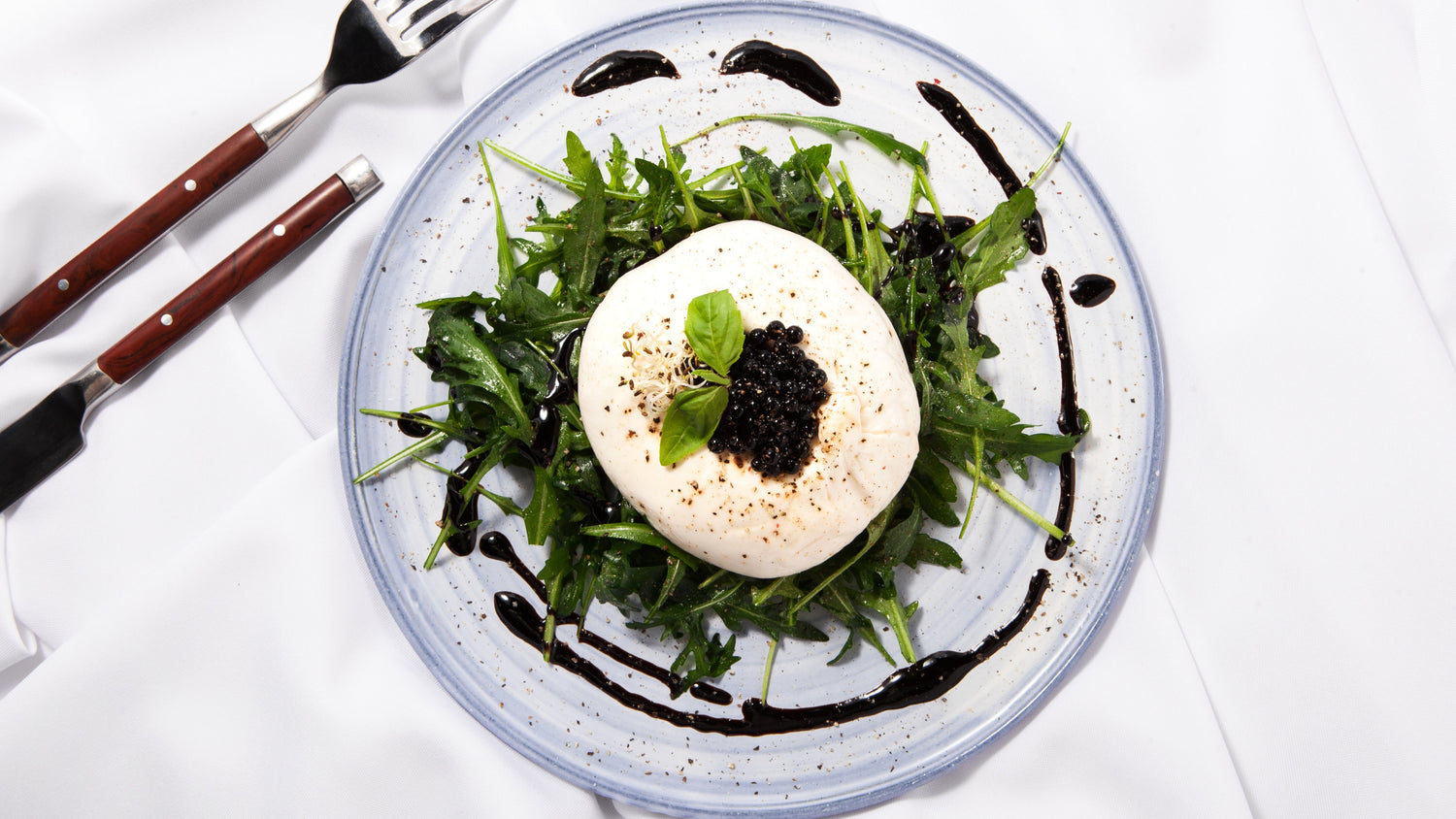 Truffle Balsamic Vinegar Pearls with Sticky Balsamic with a drizzle of balsamic around the arugula rocket, burrata, pearls and basil. On a grey plate with a knife and form next to it on a white tablecloth