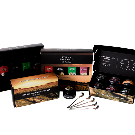 Ultimate Bundle with Sticky Balsamic including all of the products, showing two Sticky Balsamic Bottles Gift Boxes complete with product, and also two Sticky Balsamic Pearls Gift Boxes, one open and one closed, along with several spoons on a white background.
