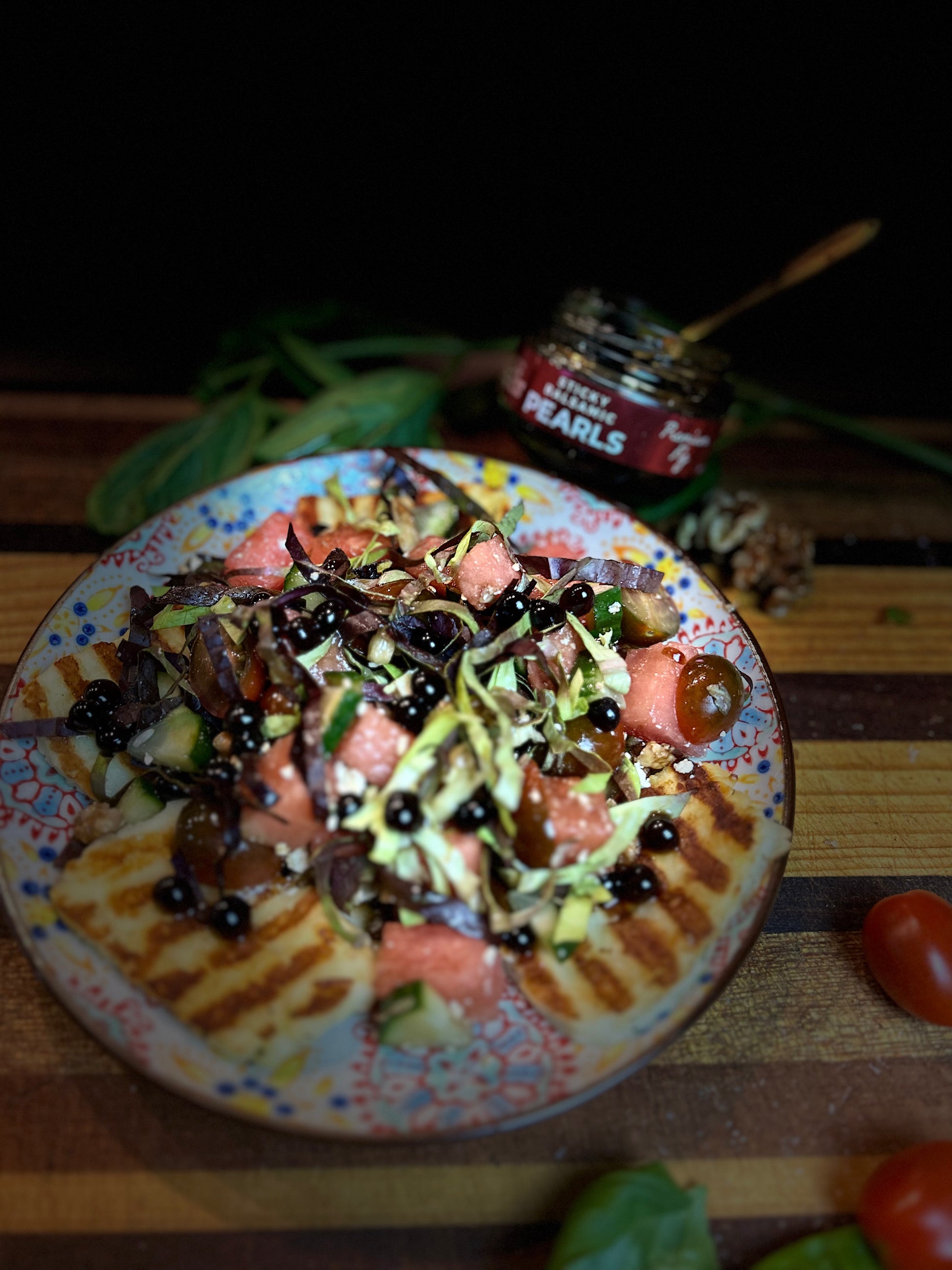 Watermelon halloumi tomato salad with pearls on a colourful plate, a jar of pearls that are opened, on a wooden chopping board with roma tomatoes and basil for decoration