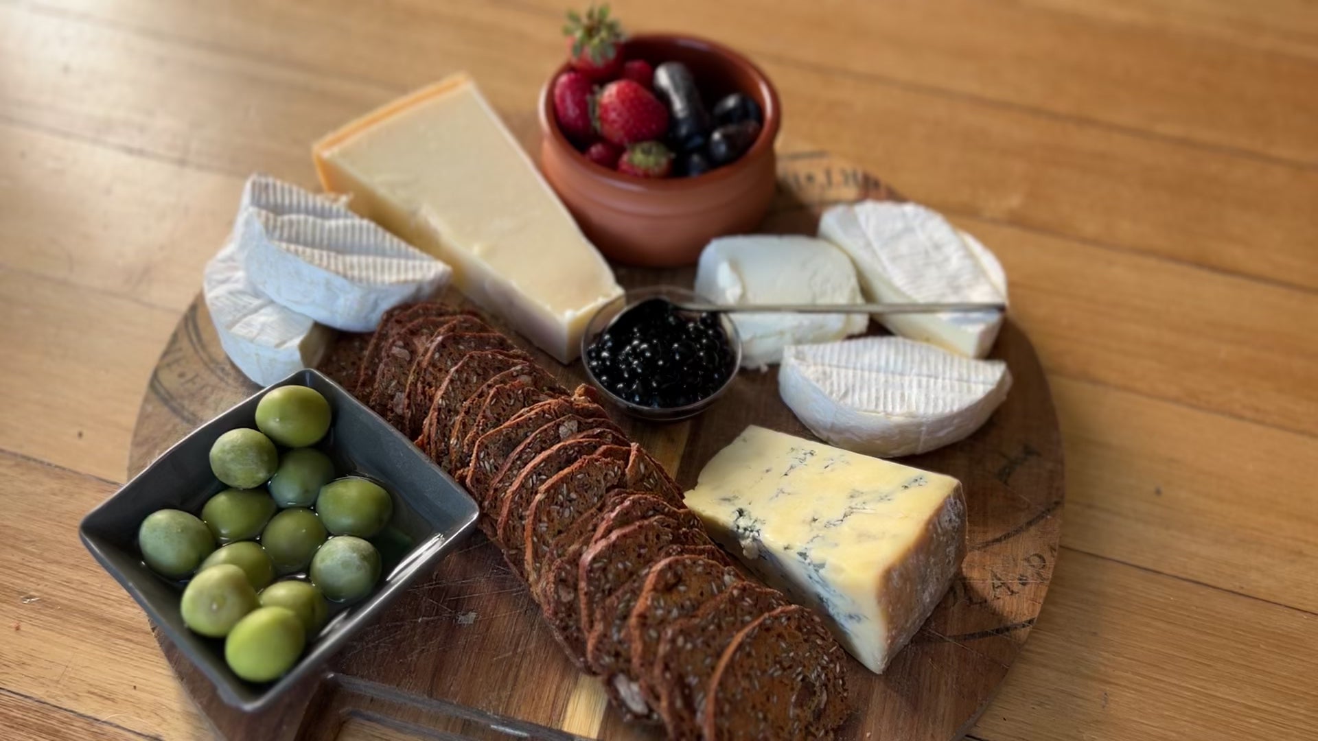 Cheese Plate and Pearls with Sticky Balsamic showing a wooden serving platter with different cheese, seedy crackers, olives and fruit, with a small bowl of Sticky Balsamic Pearls with a Sticky Balsamic Spoon in it.