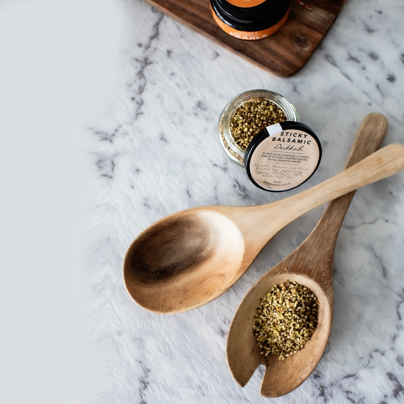 Sticky Balsamic Dukkah presented with the jar askew presented artfully with a jar of Sticky Balsamic Lemon Pearls on a wooden board, and next to serving spoons, one which has some Sticky Balsamic Dukkah in it. All of ths is on a marble background.