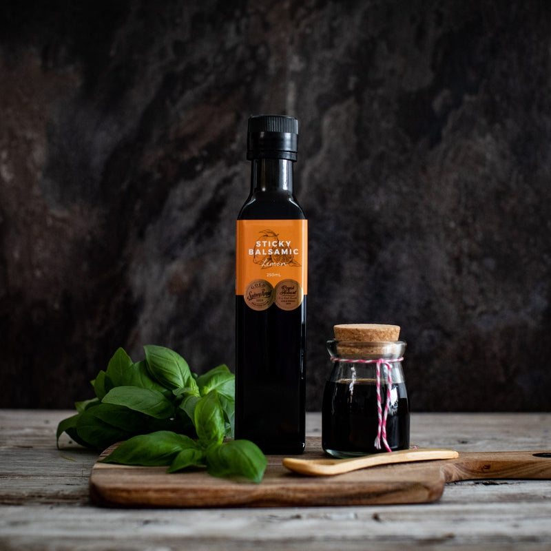 Sticky Balsamic Lemon 250ml next to a jar of balsamic on a wooden serving board next to basil and a wooden spoon. It is on a grey table with a dark, artistic background.