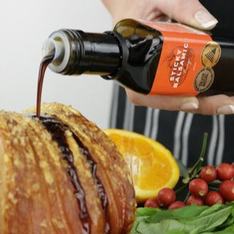 Sticky Balsamic Orange 250ml bottle being drizzled over a roast, held by a hand, in front of orange, berries and lettuce.