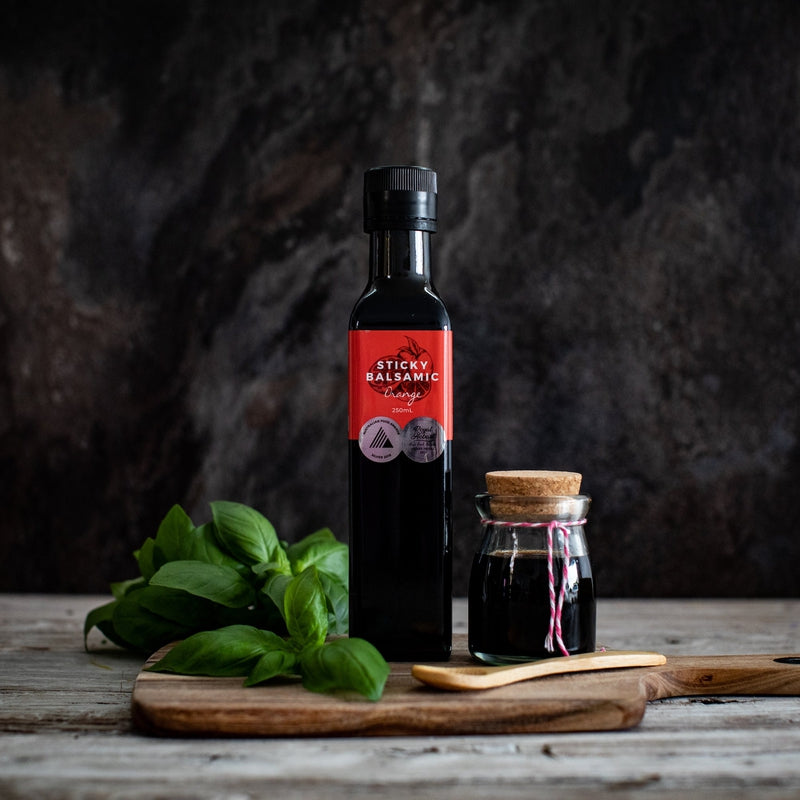 Sticky Balsamic Orange 250ml next to a jar filled with balsamic on a wooden board, next to basil and a wooden spoon, on a wooden benchtop and with a dark artistic wall behind it