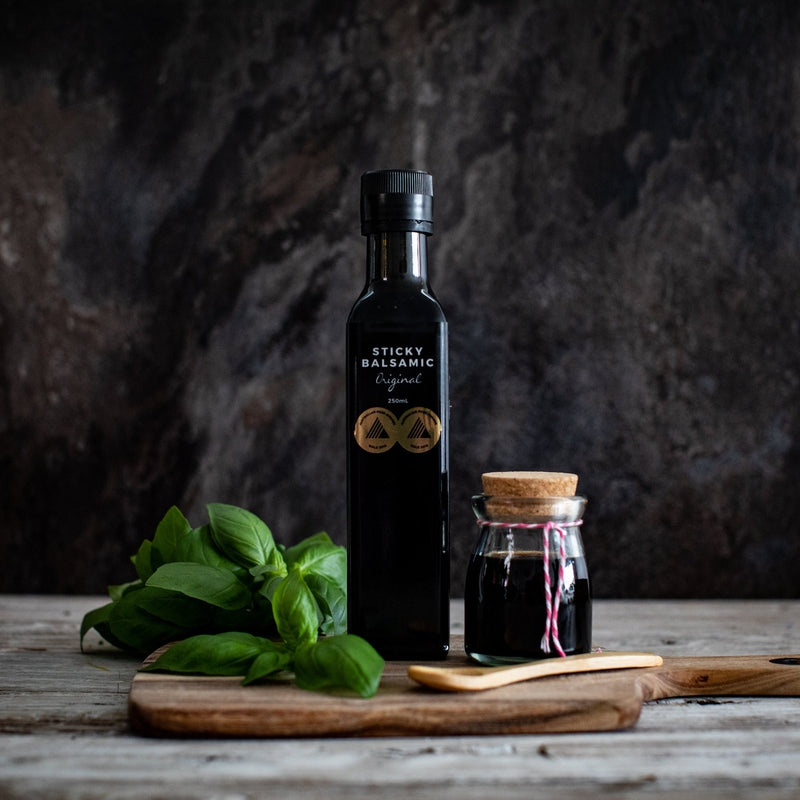 Sticky Balsamic Original 250ml next to a jar containing balsamic, on a wooden board with basil and a wooden spoon, on a wooden bench and an artistic dark background.