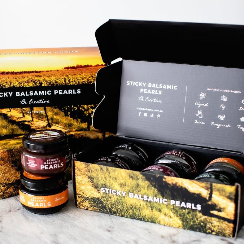 Sticky Balsamic Pearls Gift Box image with three gift boxes, two stacked on top of each other and the third open showing how the pearl jars look inside it. There are two pearls jars on top of the other next to the boxes all on a marble bench and white background.
