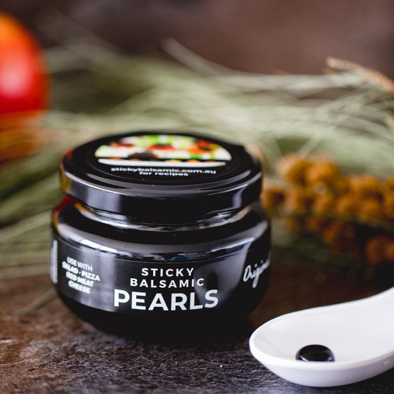 Sticky Balsamic Pearls - Original 110g net jar with a single white spoon containing a single pearl, with a background of wattle and a dark background.