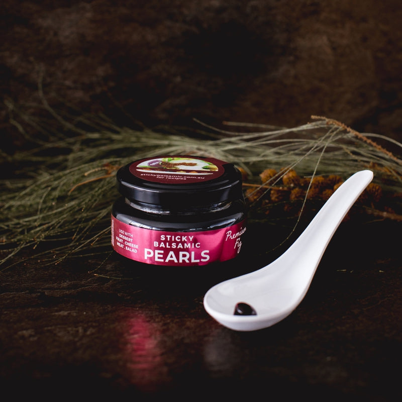 Sticky Balsamic Pearls - Premium Fig jar with a white spoon and a single pearl show its shape and form. This is with decoration of wattle and a dark artful background