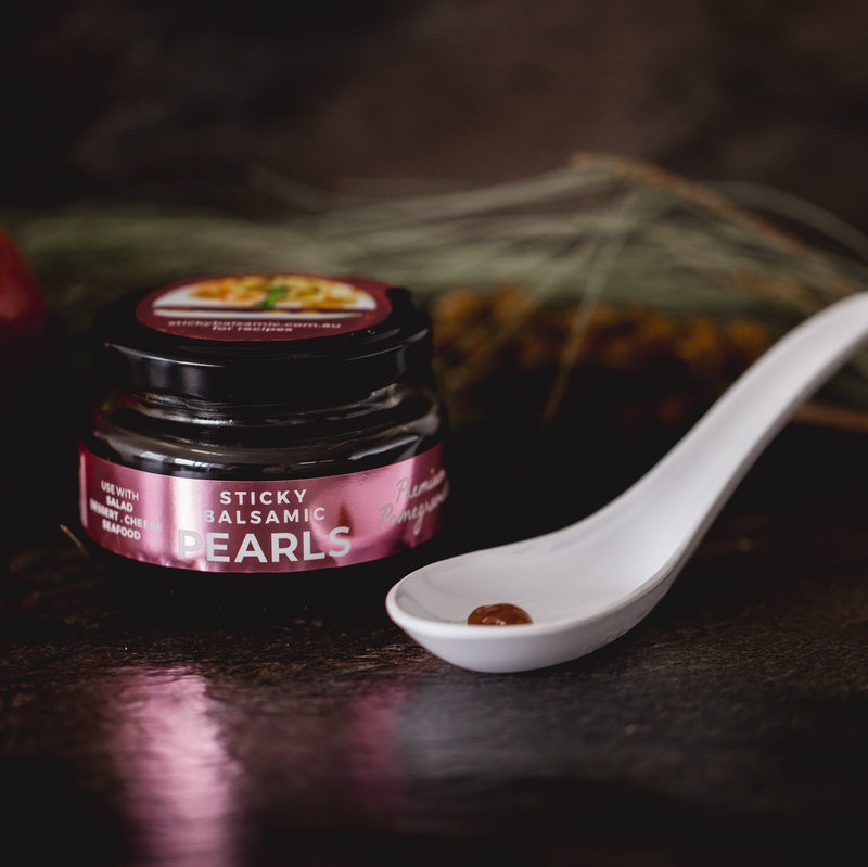 Sticky Balsamic Pearls - Premium Pomegranate 110g net jar on a dark background next to a white spoon showing and individual pearl