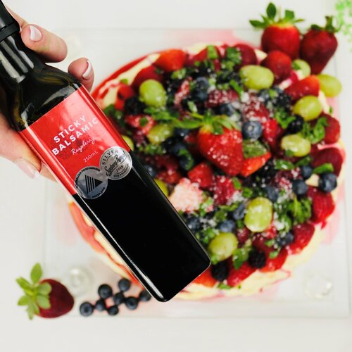 Sticky Balsamic Premium Raspberry 250ml bottle being held above a pavlova with fruit on top of it, and a delicious red syrup which contains Sticky Balsamic Premium Raspberry.