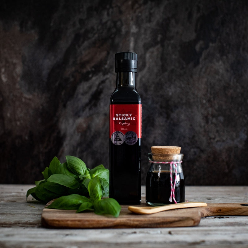 Sticky Balsamic Premium Raspberry 250ml bottle on a wooden board with basil, a wooden spoon and jar with balsamic in it. The background is dark giving it a moody artistry