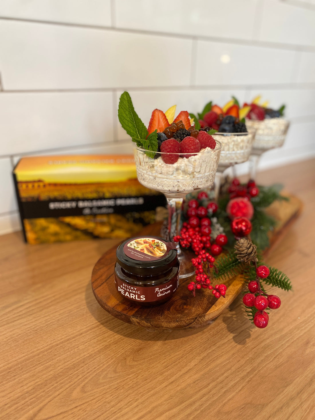 Three elegant glass bowls of overnight oats topped with fresh strawberries, raspberries, blueberries, mint leaves and Sticky Balsamic Quince Pearls with festive holiday decorations