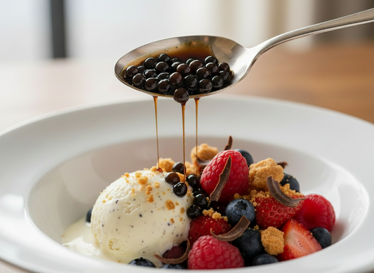 Spoonful of balsamic pearls with a glossy drizzle falling onto vanilla ice cream with fresh berries, crumbs and chocolate curls in a white bowl.
