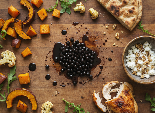 Overhead photo of dark balsamic pearls with syrup smears on a kitchen bench, surrounded by leftover roast vegetables, shredded roast chicken, flatbread, salad leaves, feta and toasted nuts.