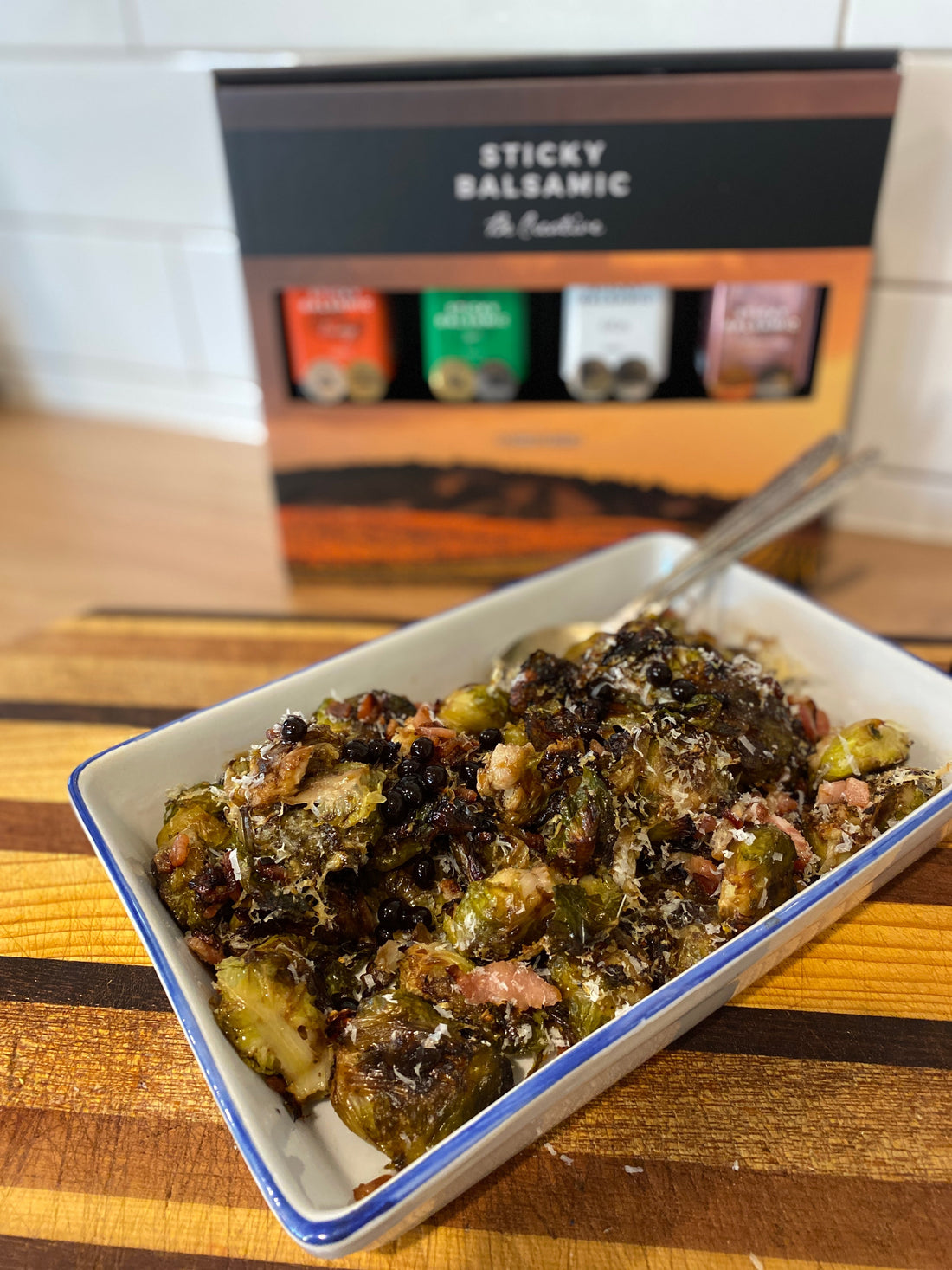 White enamel dish filled with golden crispy smashed brussels sprouts topped with bacon pieces, Sticky Balsamic Original Pearls, and grated parmesan cheese with Sticky Balsamic gift box in background