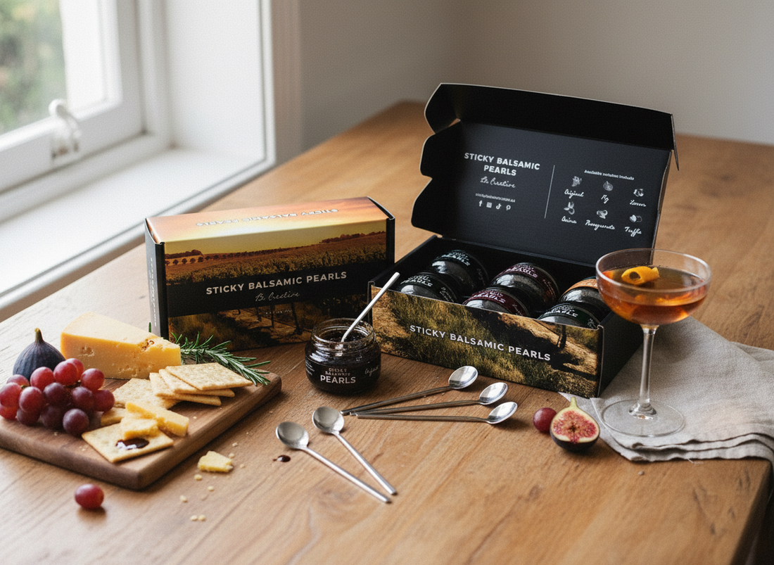 Gourmet food gift set featuring a basket of Sticky Balsamic Pearls with assorted cheeses, dried fruit, nuts, crackers, chocolate, and a cocktail garnished with balsamic pearls on a rustic wooden table.