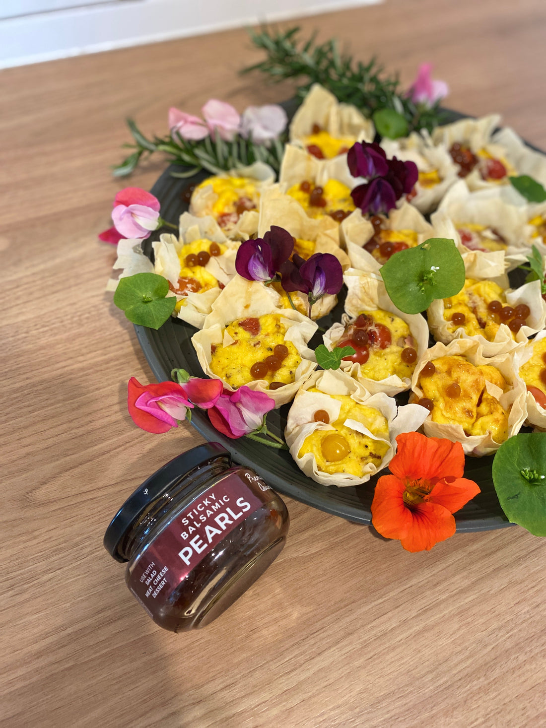 Platter of golden mini filo quiches topped with Sticky Balsamic Quince Pearls and garnished with purple and orange edible flowers and nasturtium leaves on black serving board