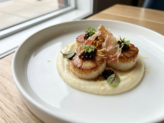 Pan-seared scallops served on creamy cauliflower purée, garnished with Sticky Balsamic Lemon Pearls and delicate balsamic drizzle, crispy prosciutto shards, lemon zest, and micro herbs. Elegant, restaurant-style plating with a modern, gourmet touch.