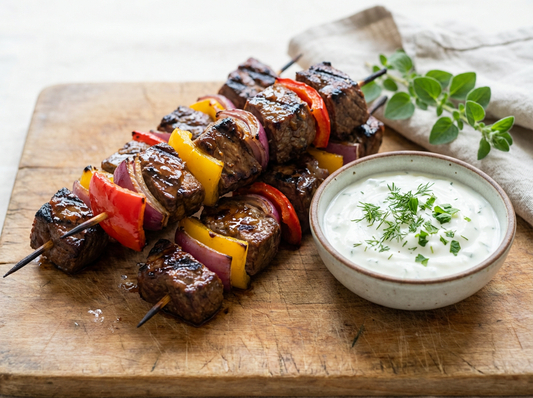 Grilled beef skewers glazed with Sticky Balsamic Black Garlic, featuring juicy, charred beef cubes, vibrant red and yellow capsicum, and red onion, arranged on a rustic wooden board. 