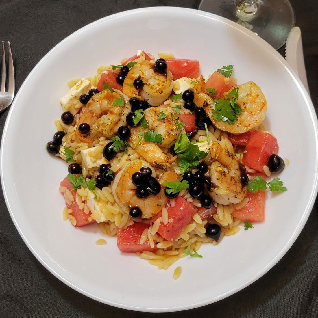 Watermelon orzo salad with grilled shrimp, feta cheese, fresh cilantro, and Fig Sticky Balsamic Pearls served on a white plate with wine glass