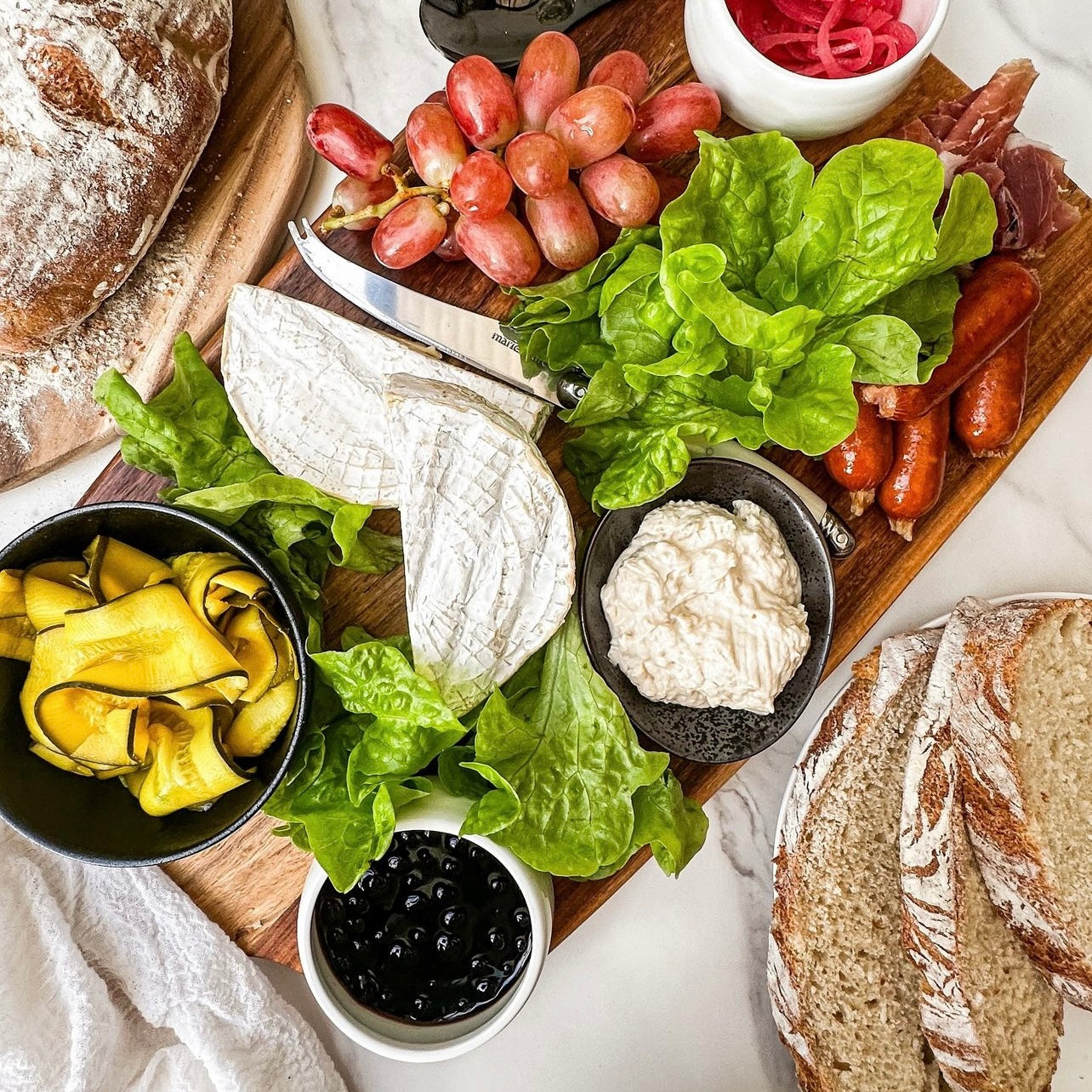 Cheese Board my family plate with Sticky Balsamic Pearls, pickled zucchini, deli meatsdips, lettuce, cheese and bread on a wooden board on a marble bench.