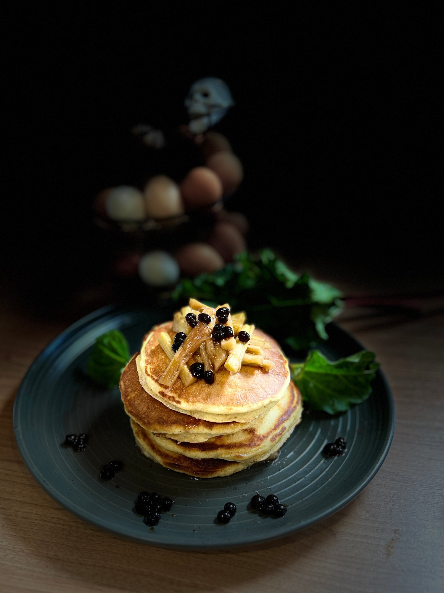 Stack of pancakes topped with sliced fruit and small dark berries on a dark plate with greens in the background