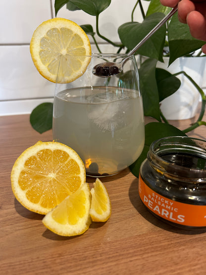 Lemon cocktail with soda water and limoncello presented with ice in a glass cup and a slice of lemon to decorate the cup. Lemon is cut and in front of the cup and is next to an open jar of Sticky Balsamic Pearls. A plant is in the background which includes subway tiles and a wooden benchtop.