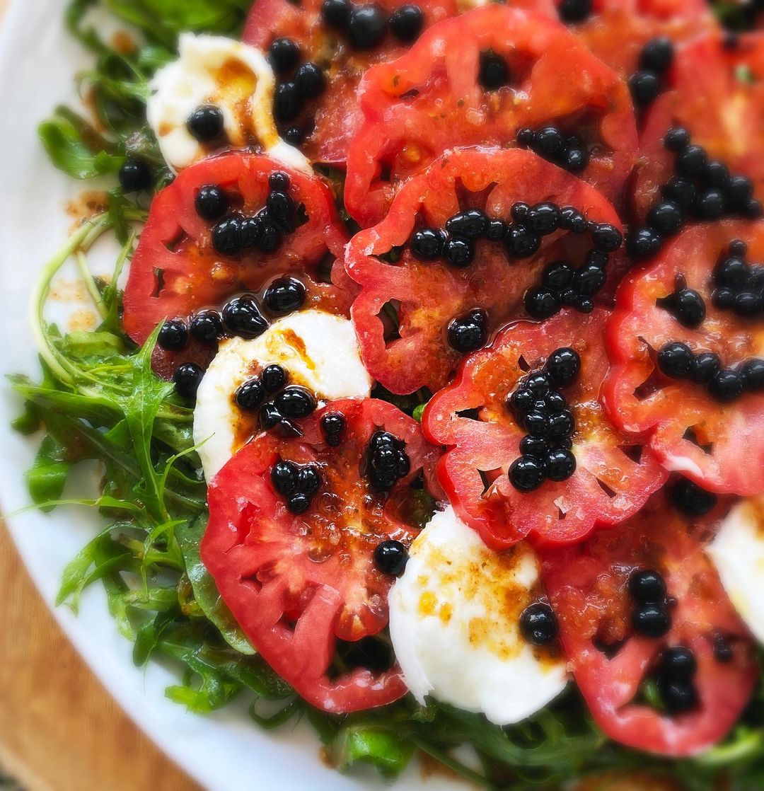 Caprese Salad with heirloom tomatoes, sticky balsamic pearls, mozzarella, rocket/arugula on a white plate.