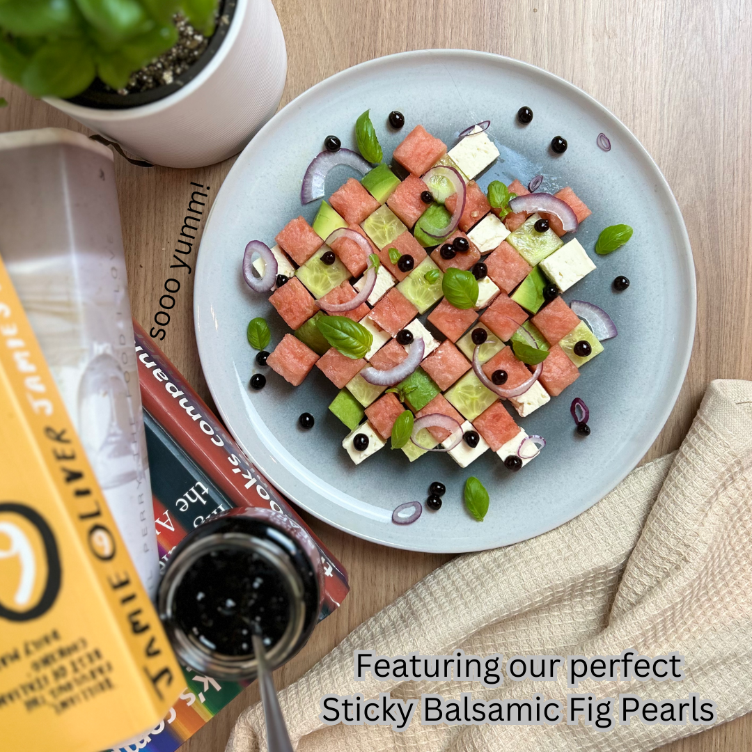 Watermelon and Feta Checkerboard Salad with watermelon, feta, avocado, cucumber, basil and red onion on a plate. There are books in a stack with a jar of Sticky Balsamic Pearls on it also, including a basil plant on a wooden bench.