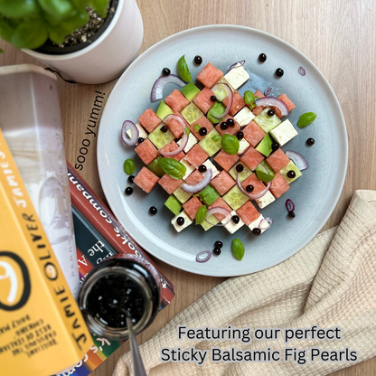 Watermelon and Feta Checkerboard Salad with watermelon, feta, avocado, cucumber, basil and red onion on a plate. There are books in a stack with a jar of Sticky Balsamic Pearls on it also, including a basil plant on a wooden bench.