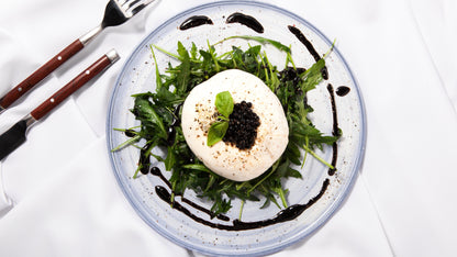Truffle Balsamic Vinegar Pearls with Sticky Balsamic with a drizzle of balsamic around the arugula  rocket, burrata, pearls and basil. On a grey plate with a knife and form next to it on a white tablecloth