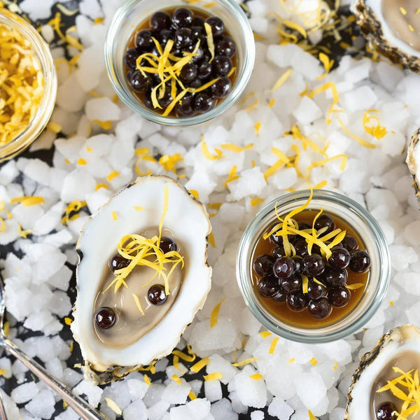Oysters on a bed of ice with lemon zest garnished with Sticky Balsamic pearls, with a jar of sticky balsamic pearls.