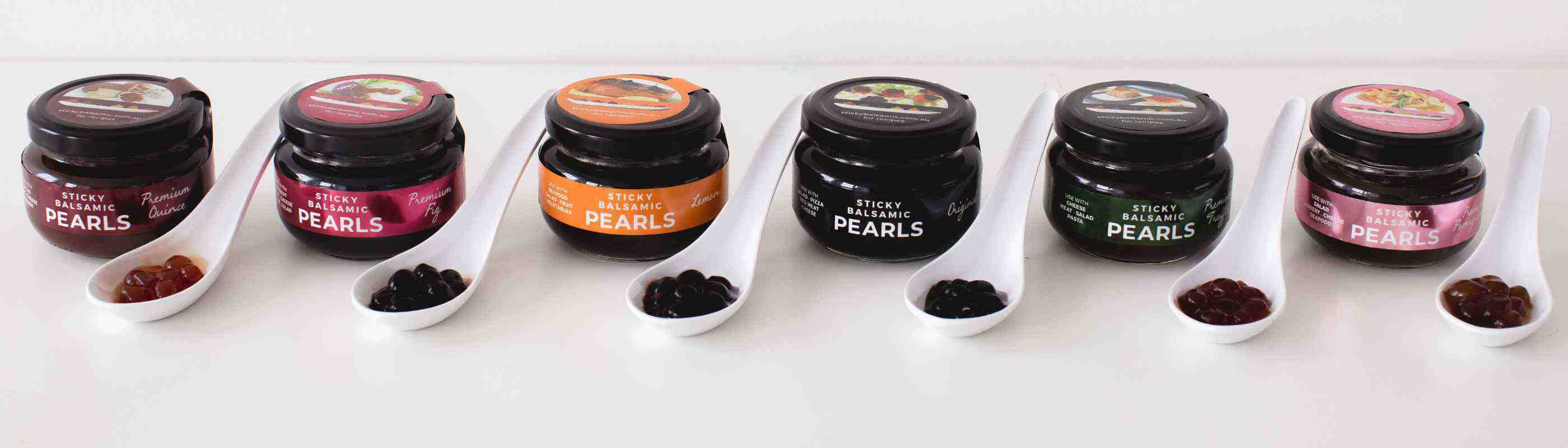 Six small glass jars of colorful caviar-like pearls with matching spoons arranged in a row on a white surface
