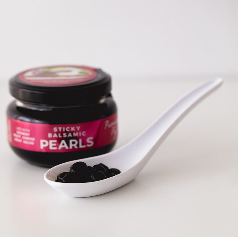 White spoon filled with dark sticky pearls beside small jar of sticky balsamic premium fig pearls